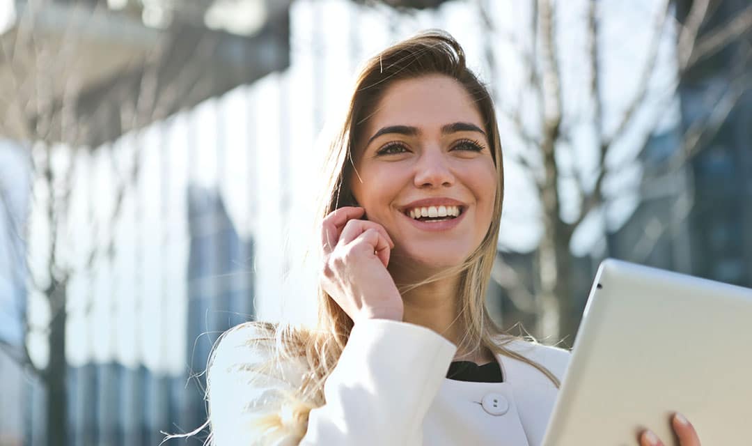 Woman with long hair and straight teeth talking on the phone while holding a tablet